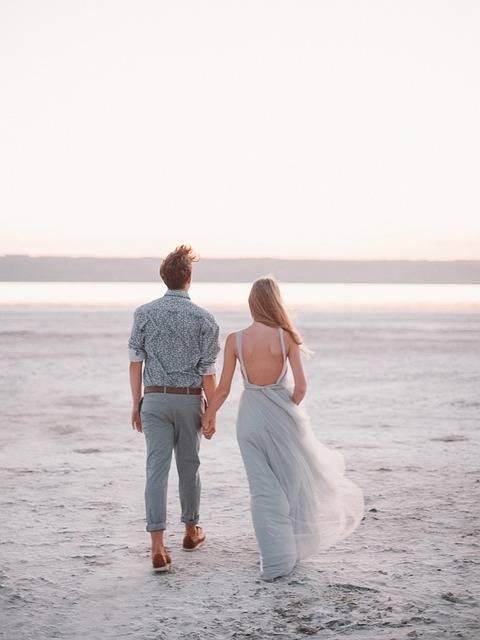 Pareja caminando de la mano por una playa desierta al atardecer, con el mar y el cielo en tonos suaves de rosa y azul. La mujer lleva un vestido ligero y el hombre una camisa de estampado, ambos disfrutando del momento juntos.