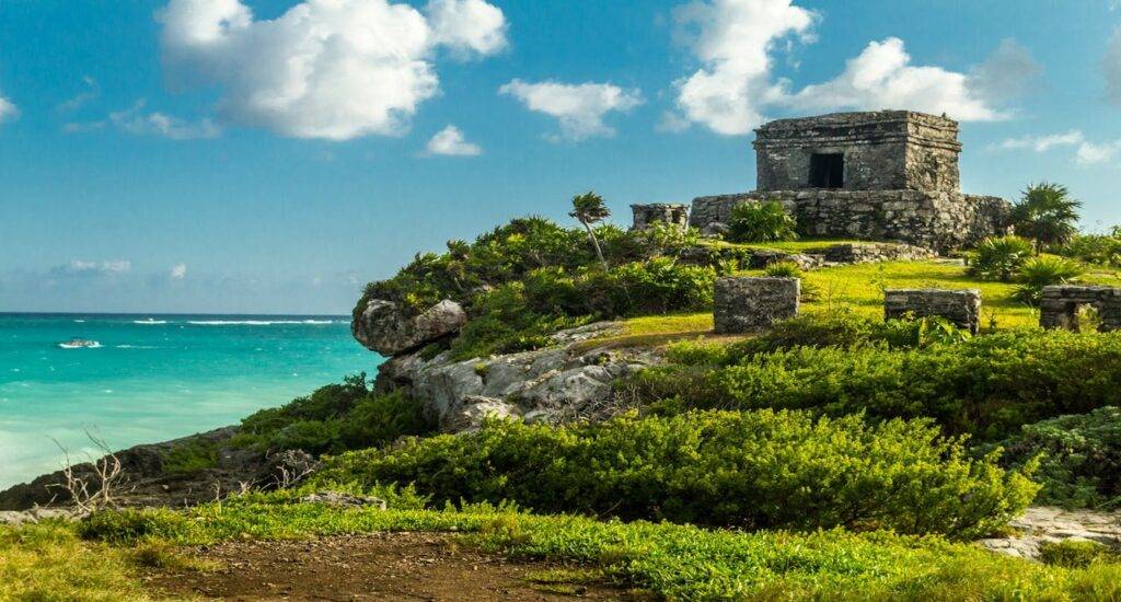 Ruinas mayas sobre un acantilado, con vistas al mar turquesa y un cielo parcialmente nublado. La vegetación verde rodea el sitio arqueológico.