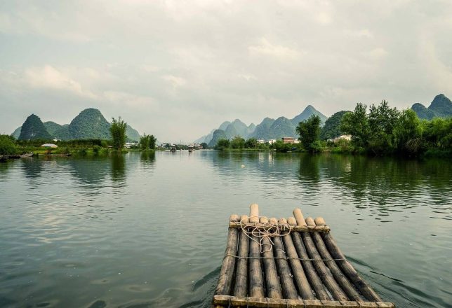 Una balsa de bambú flota en un río rodeado de montañas verdes y un cielo nublado. La escena transmite tranquilidad y conexión con la naturaleza.